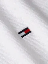 Chomba blanca de corte slim con cuello mao, tapeta de tres botones y logo de Tommy Hilfiger bordado en el pecho. El cuello presenta una banda con los colores característicos de la marca: azul marino, blanco y rojo.