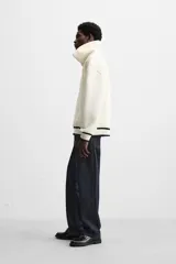 Sudadera color crudo de tejido técnico con cuello alto y cierre de cremallera. Tiene manga larga, corte relaxed fit, bolsillo tipo canguro y rib en puños y bajo con rayas negras.
