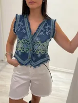 Chaleco corto de denim azul con bordados florales en blanco y marrón, detalle desflecado en escote, sisas y ruedo. Escote en V y cierre frontal con botones. Estilo boho chic.