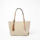Bolso tipo shopper color beige con textura de espiga y detalles en cuero color camel. Tiene doble asa de hombro y un dije de borla en uno de los laterales.