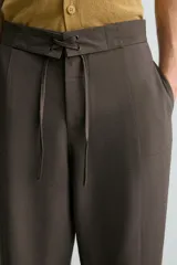 Pantalón marrón de corte ancho, con cintura ajustable con cordón, bolsillos delanteros y traseros tipo parche, detalle de costuras verticales en el frente y cierre frontal con cremallera y gancho metálico oculto.