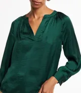 Blusa verde esmeralda de tela satinada, con escote en V, mangas largas con puños abotonados y espalda fruncida.
