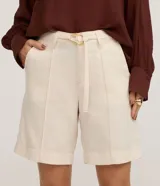 Bermuda de cintura mediana color beige, confeccionada en sarga, con trabillas, cierre tradicional, bolsillos y frisos delanteros. Incluye cinturón de tejido para lazo con hebilla metalizada.