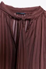 Blusa color marrón con rayas verticales, cuello subido con lazo, escote pico, mangas largas abullonadas con elástico en los puños y cierre frontal con botones forrados.