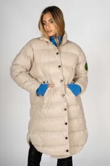 Campera puffer larga color beige, con cuello alto y cierre frontal con botones a presión. Tiene bolsillos laterales y un parche con el logo de la marca en la manga izquierda.