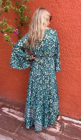 Vestido largo estilo cruzado (wrap dress) de manga larga acampanada, con estampado floral en tonos verde petróleo, amarillo pálido y celeste. Presenta un ribete decorativo en el escote y el borde del cruce.