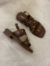 Sandalias bajas de cuero marrón con tiras cruzadas y apliques de tachas doradas.