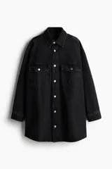 Camisa oversized de denim negro, con cuello clásico, cierre frontal con botones metálicos, dos bolsillos con solapa en el pecho, mangas largas con puños abotonados y bajo redondeado.