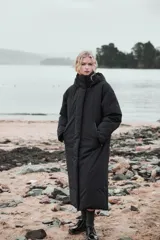 Anorak largo negro resistente al agua y al viento, con aislamiento térmico, cuello alto con capucha ajustable, bolsillos con cremallera, acolchado interior, tirantes elásticos y cremalleras laterales en el bajo.