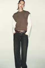 Prenda de manga larga blanca con un chaleco de tejido de punto azul marino superpuesto, creando un efecto de dos piezas. El chaleco es de cuello redondo y corte recto.