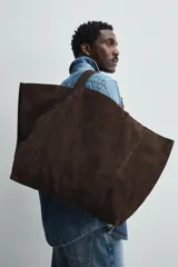 Bolso tipo shopper marrón oscuro, fabricado en cuero con acabado serraje. Tiene un compartimento principal con cierre magnético y un bolsillo interior con cremallera. Cuenta con dos asas de mano de cuero.