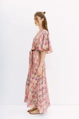 Vestido largo cruzado con estampado floral en tonos rosa, lila y dorado. Tiene mangas cortas amplias, escote en V y cintura elastizada con lazo.