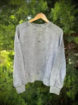 Buzo de algodón y poliéster, color gris lavado con efecto acid wash. Presenta cuello redondo con detalle de V en el centro, corte corto y calce suelto. Tiene un pequeño bordado de dos hojas estilizadas en el pecho.