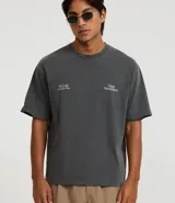 Remera de algodón de corte boxy, color gris oscuro con efecto desteñido/vintage. Presenta un estampado minimalista en blanco en el pecho con tres círculos y texto a la derecha que dice "United Victory Match".