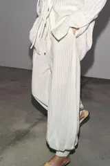 Pantalón de tiro medio color crudo con rayas verticales, confeccionado en hilatura con lino. Tiene bolsillos laterales ocultos en costura y de vivo en espalda, detalle de pliegues y bajo acabado en puño.