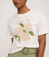 Remera blanca de algodón, con cuello redondo y manga corta. Presenta un estampado frontal con la frase "SOY FLOR. SOY LATINA" y un diseño floral con flores en relieve y hojas bordadas.