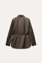 Camisa marrón de Zara Woman Collection, confeccionada en hilatura con viscosa. Tiene cuello solapa, manga larga con puño, bolsillo de plastrón en el pecho, cinturón ajustable con lazadas y bajo asimétrico. Se cierra con botones frontales.