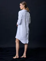 Vestido camisero blanco de algodón con cuello solapa, escote en V y mangas 3/4 regulables. Presenta bolsillos laterales verticales y abertura con botones en la espalda.