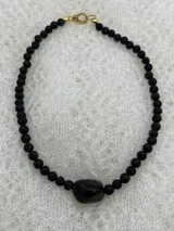Collar corto de cuentas redondas de piedra volcánica negra, con cuenta central irregular de piedra negra pulida y cierre de gancho dorado.
