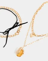 Set de tres collares dorados con diferentes tipos de cadenas y dijes: gargantilla de terciopelo negro con lazo, cadena con dije de corazón y cadena con relicario ovalado con diseño floral.