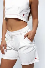 Short blanco de gabardina con pespuntes rojos, tiro medio/alto, tablones delanteros, bolsillos delanteros y traseros estilo jean. Presenta un logo bordado en rojo en la pierna derecha.