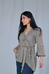 Blusa color gris beige con escote cruzado en V y volados en el frente. Tiene mangas largas con puños elásticos y corte a la cintura con lazo lateral.