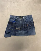 Minifalda de denim azul oscuro, confeccionada con retazos de jeans, con tiro bajo y borde inferior deshilachado. Presenta detalles de costuras y bolsillos de jeans, incluyendo un parche con la inscripción "RAPSODIA" en el bolsillo delantero.