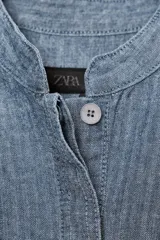 Camisa de denim azul con cuello redondo, manga corta abullonada con pliegues y bajo abullonado con pliegues. Cierre frontal con botones ocultos por solapa.