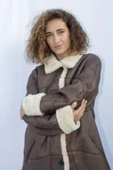 Abrigo gamulán marrón con cuello, solapa, puños y borde frontal de piel sintética blanca. Tiene cierre con botones y bolsillos laterales.