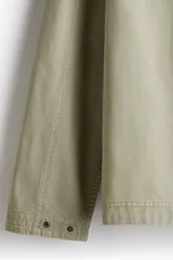 Sobrecamisa color arena de lona gruesa de algodón, corte recto, con cuello clásico, cierre frontal con botones a presión ocultos, canesú en la espalda, dos bolsillos superiores con solapa y botón a presión, bolsillos laterales discretos, mangas largas con puños abotonados y forro interno.