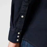Camisa de denim azul oscuro con cuello americano, cierre con botones a presión blancos, dos bolsillos frontales con solapa y logo de la marca bordado en el bolsillo izquierdo.