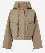 Campera corta color marrón claro, confeccionada en sarga de algodón y poliamida, con cuello alto, capucha y bolsillos delanteros con solapa. Tiene cierre frontal con botones a presión y mangas largas con puños elásticos.