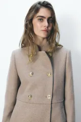 Abrigo color beige confeccionado en lana. Tiene cuello subido, mangas largas con hombreras, forro interior combinado y cierre frontal cruzado con botones metálicos.