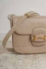 Bandolera color beige con textura símil reptil, cierre con broche metálico dorado y correa larga regulable.