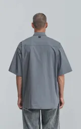 Camisa gris de manga corta confeccionada en poplin 100% algodón, con silueta oversize y dos bolsillos delanteros, uno con solapa y otro con ojal y botón.