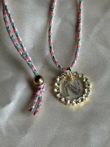 Collar con dije circular dorado con strass y letra 'A' en el centro, con cordón trenzado multicolor y cierre ajustable.