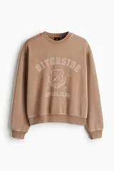 Buzo de tejido pesado de algodón con efecto lavado, color marrón topo. Presenta un diseño estampado en el frente con el texto "RIVERSIDE SOCIAL CLUB" y un escudo con un remo. Tiene cuello redondo, hombros caídos y puños y bajo acanalados.