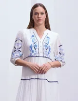 Vestido midi blanco con bordados florales azules en el escote, mangas y cintura. Tiene cuello en V, mangas 3/4 con botón y falda escalonada con detalles de ribetes azules.