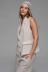 Chaleco sastrero sin mangas color beige con rayas verticales blancas, cuello solapa, botones delanteros y bolsillos con tapa.