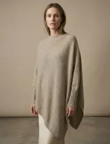 Poncho de tejido lurex color beige jaspeado, con cuello redondo y mangas largas con puños acanalados. Presenta un corte asimétrico con caída pronunciada en los laterales.