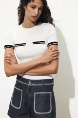 Camiseta blanca de corte recto con cuello redondo y mangas cortas. Presenta dos falsos bolsillos en el delantero con detalle de fornitura a contraste en negro y dorado.