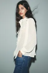 Camisa blanca semitransparente con cuello subido, manga larga con puños, pliegues y volantes combinados. Cierre frontal con botones.