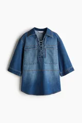 Camisa popover de denim azul con costuras en color contrastante, corte oversized, hombros caídos, cuello abierto con detalle de cordón, bolsillos grandes superiores, mangas al codo y bajo levemente redondeado.