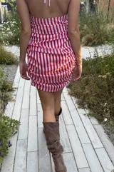 Vestido corto de gasa con estampado de rayas verticales rojas y blancas. Presenta un escote pronunciado en V, diseño halter y drapeado lateral en la cadera con un lazo largo ajustable. Incluye un lazo adicional a juego para usar como accesorio.