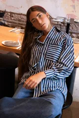 Camisa de rayas verticales azules y blancas, con cuello camisero, manga larga y puños abotonados. Presenta un bolsillo en el pecho con ribete rojo y un detalle de ribete rojo en el cuello y la tapeta de botones.