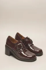 Zapato de cuero vacuno charol negro, con punta redondeada y detalle de perforado en la capellada. Tiene una tira con hebilla metálica y un taco cuadrado de 4 cm.