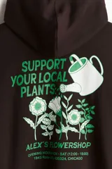 Buzo canguro marrón con capucha cruzada, hombros caídos, bolsillo canguro y puños y bajo acanalados. Estampado frontal con texto en verde. Corte holgado.