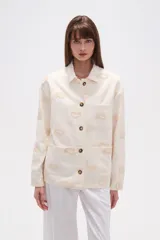 Campera de algodón color crudo, estilo camisa o chaqueta ligera, con bordados en forma de paisley en tono beige claro. Posee cuello camisero, cierre frontal con botones marrones y bolsillos delanteros.