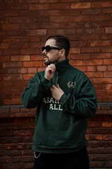 Buzo verde oscuro de algodón con cierre medio frontal, cuello alto y corte oversize. Estampado frontal con la frase "Against All" en color crema y detalles bordados.