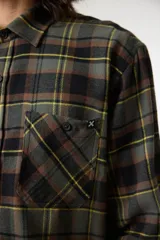 Camisa de manga larga con cuello camisero y botones frontales. Presenta un patrón de cuadros en tonos verde oliva, negro, marrón y amarillo. Cuenta con dos bolsillos frontales con solapa.